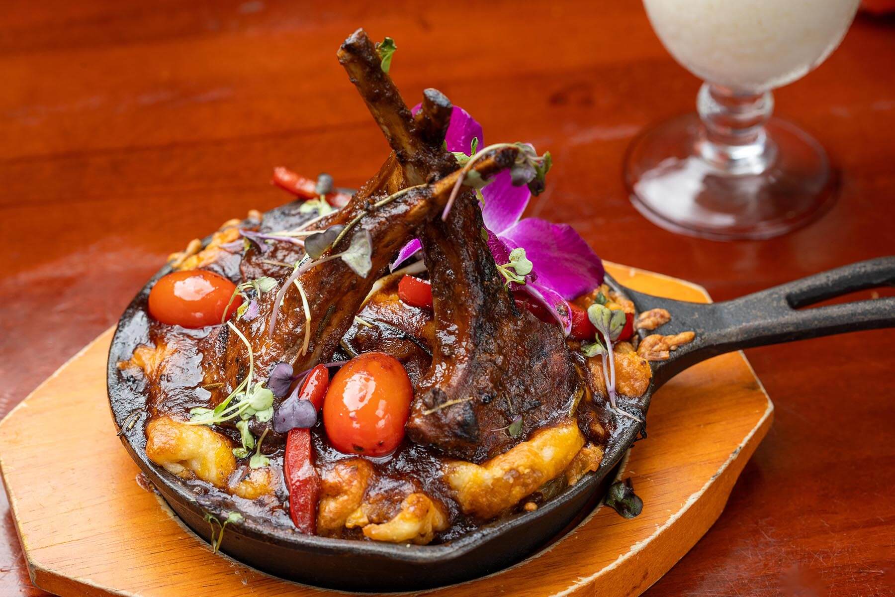 Skillet with grilled lamb chops, cherry tomatoes, red peppers, melted cheese, garnished with microgreens and a purple flower on a wooden board.