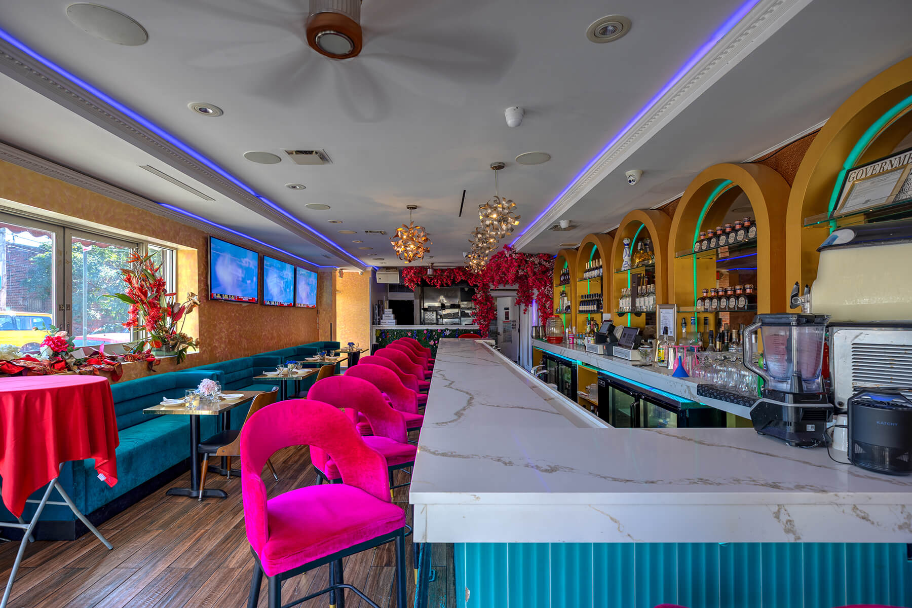 Modern bar interior with white marble counter, bright pink bar stools, teal seating along wall, and decorative red flowers.