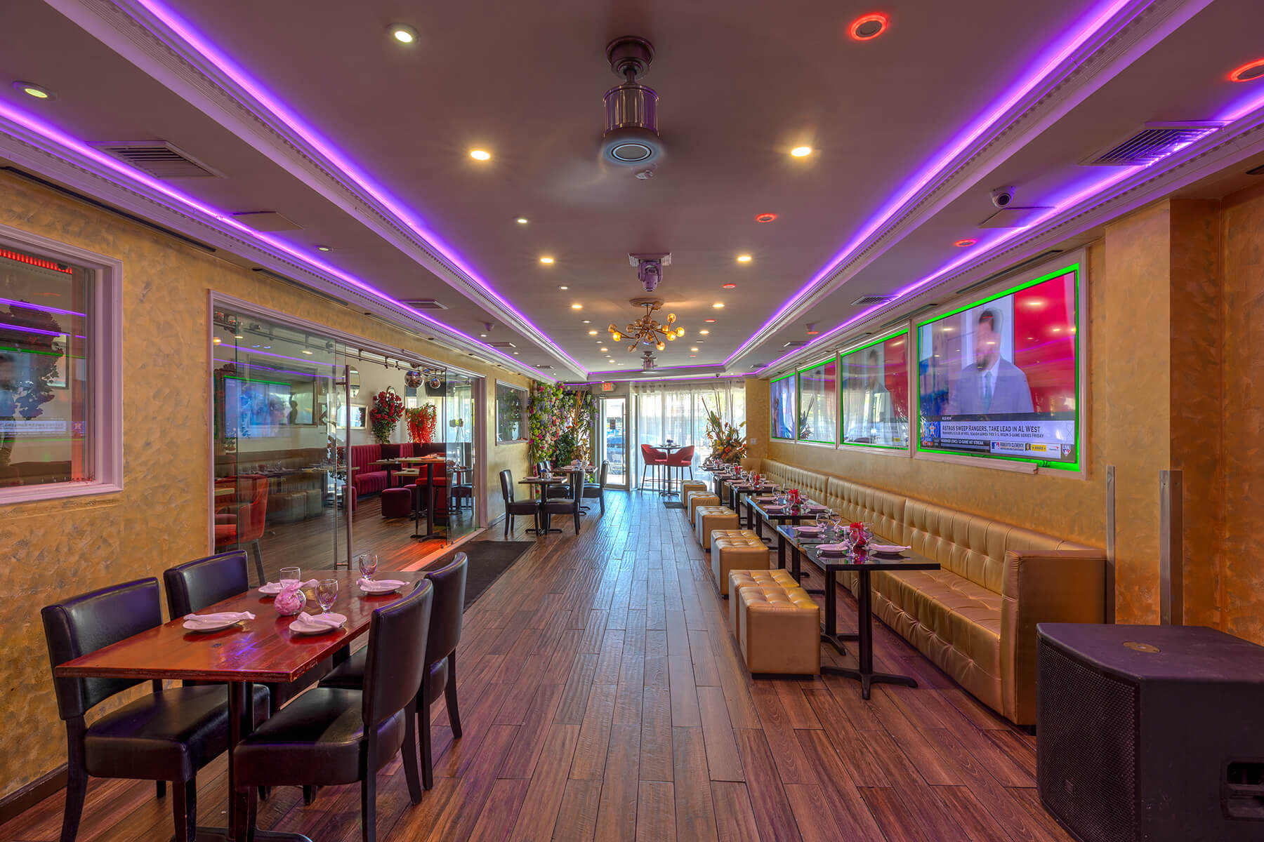 Modern restaurant interior with wood flooring, purple LED ceiling lights, multiple tables with chairs on the left, and a long tan bench with small tables and stools on the right.