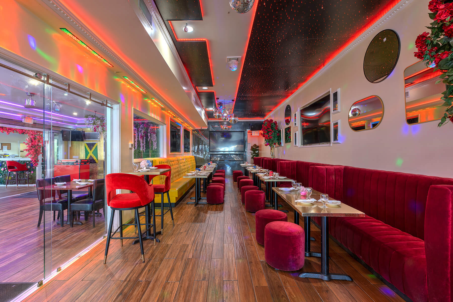 Modern restaurant interior with red and yellow velvet seating, wooden floors, mirrored walls, and colorful ambient lighting.