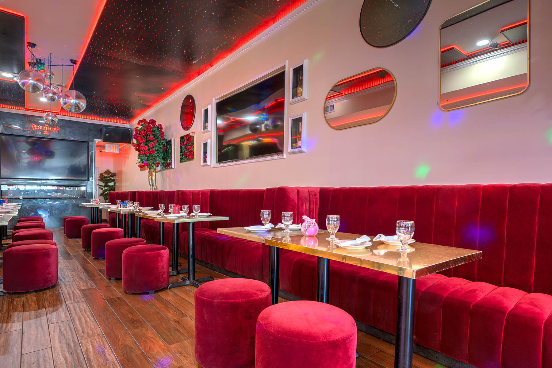 Modern restaurant interior with red velvet banquette seating, matching round stools, gold tables, mirrors on the wall, and ambient red lighting.