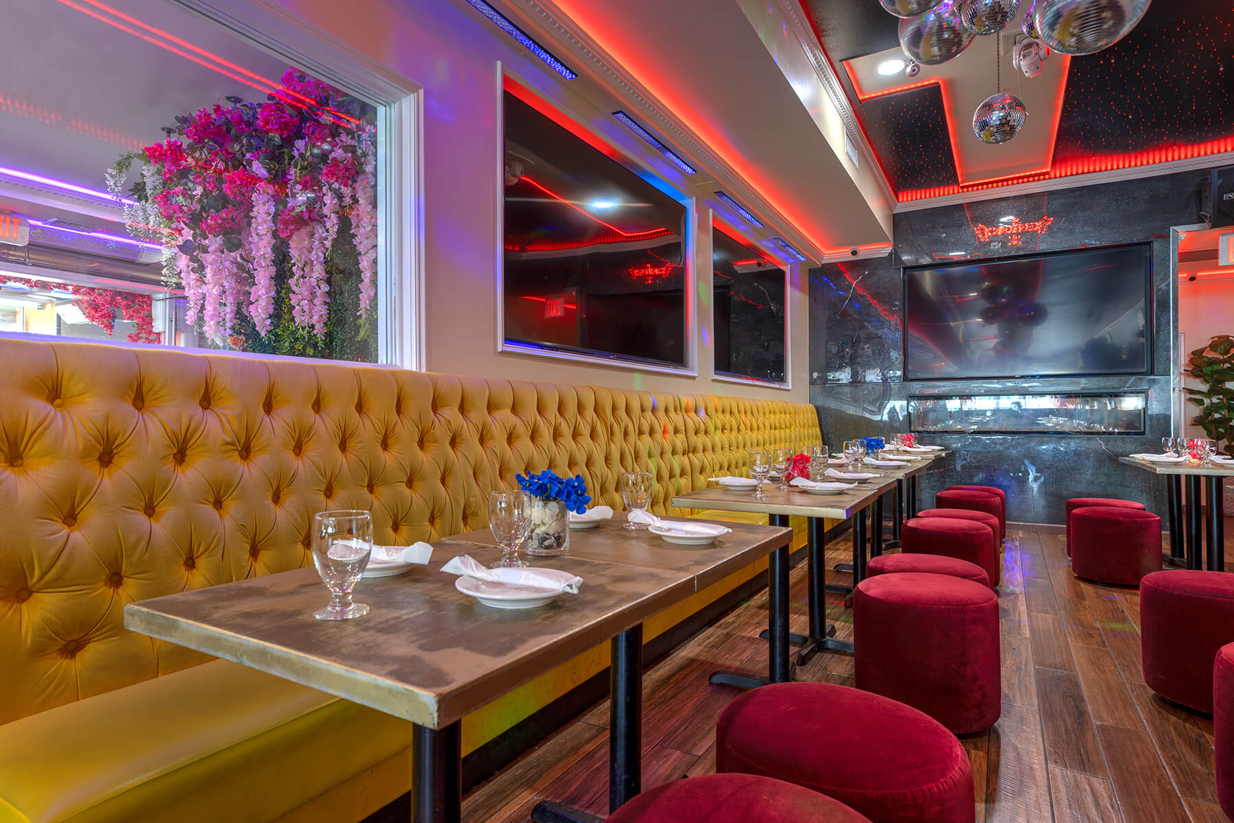 Modern restaurant interior with mustard yellow tufted bench seating, red velvet stools, wooden tables set with glassware and napkins, floral window decor, and large flat-screen TVs on dark marble walls under red ambient lighting.