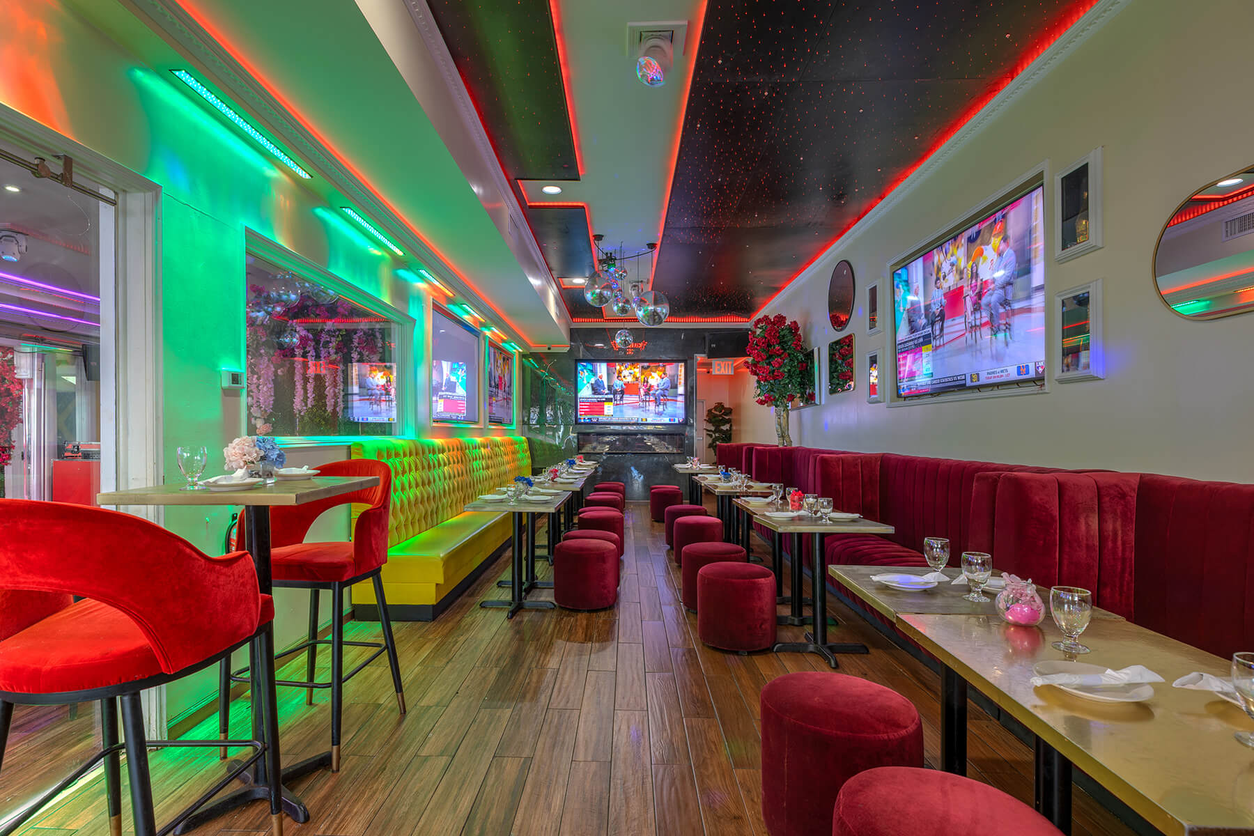 Interior of a modern restaurant with red velvet chairs and stools, yellow tufted bench seating, colorful neon lighting, multiple wall-mounted TVs, and wood flooring.