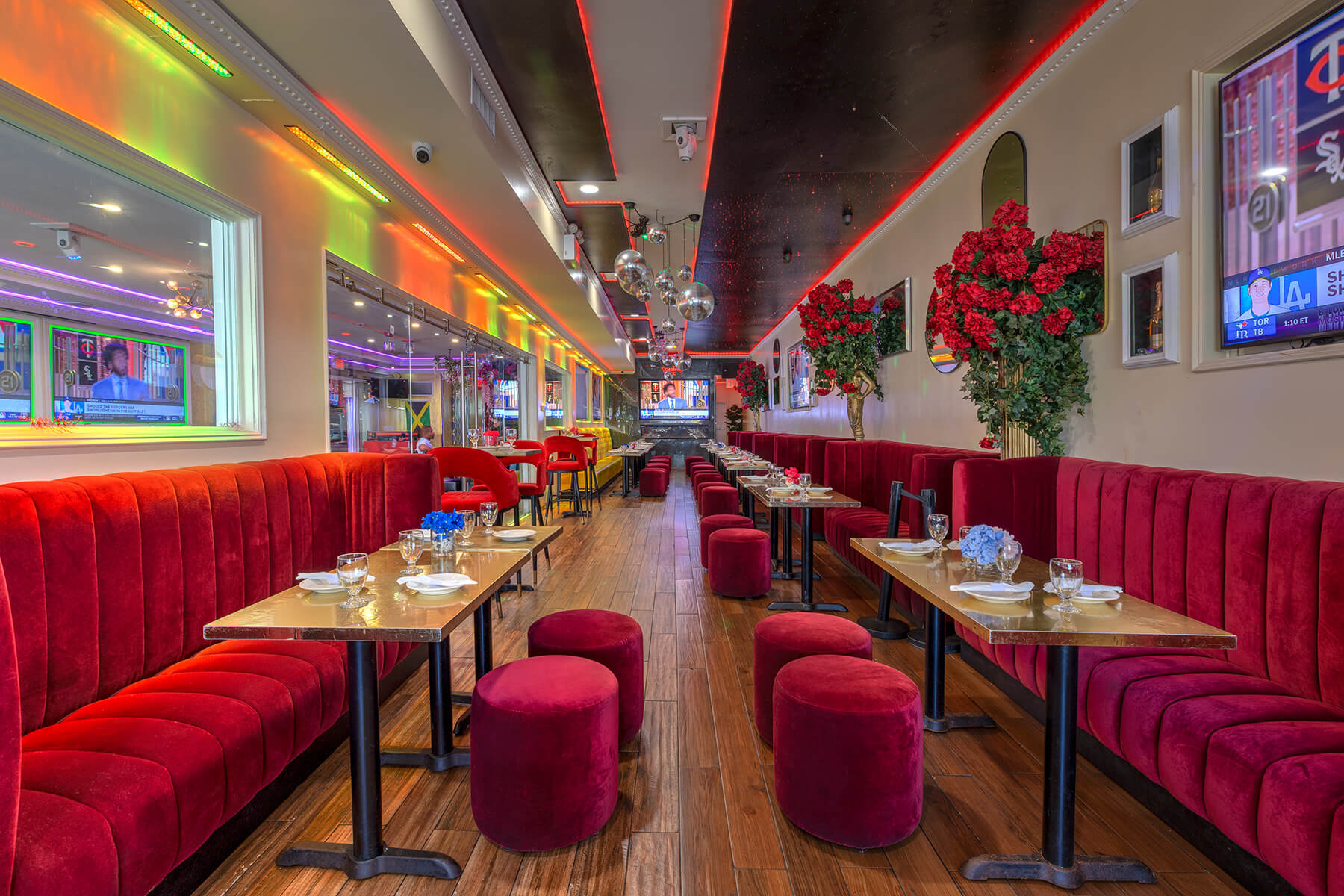 Modern restaurant interior with red velvet banquettes, round stools, wooden floors, colorful LED lighting, and decorative red flowers.