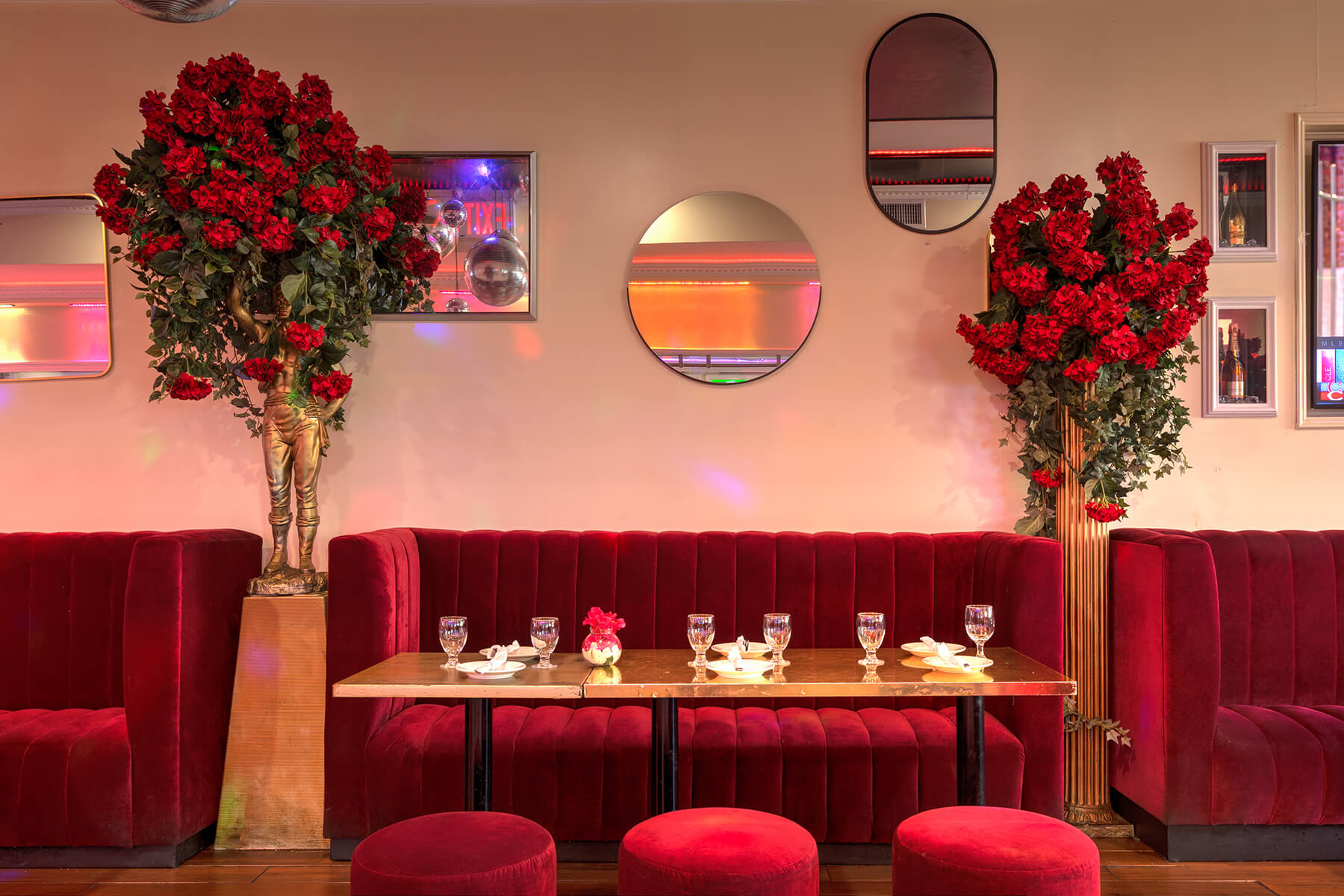 Luxurious restaurant seating with red velvet booths and stools, a wooden table set with glassware, and tall floral arrangements with red flowers on either side.