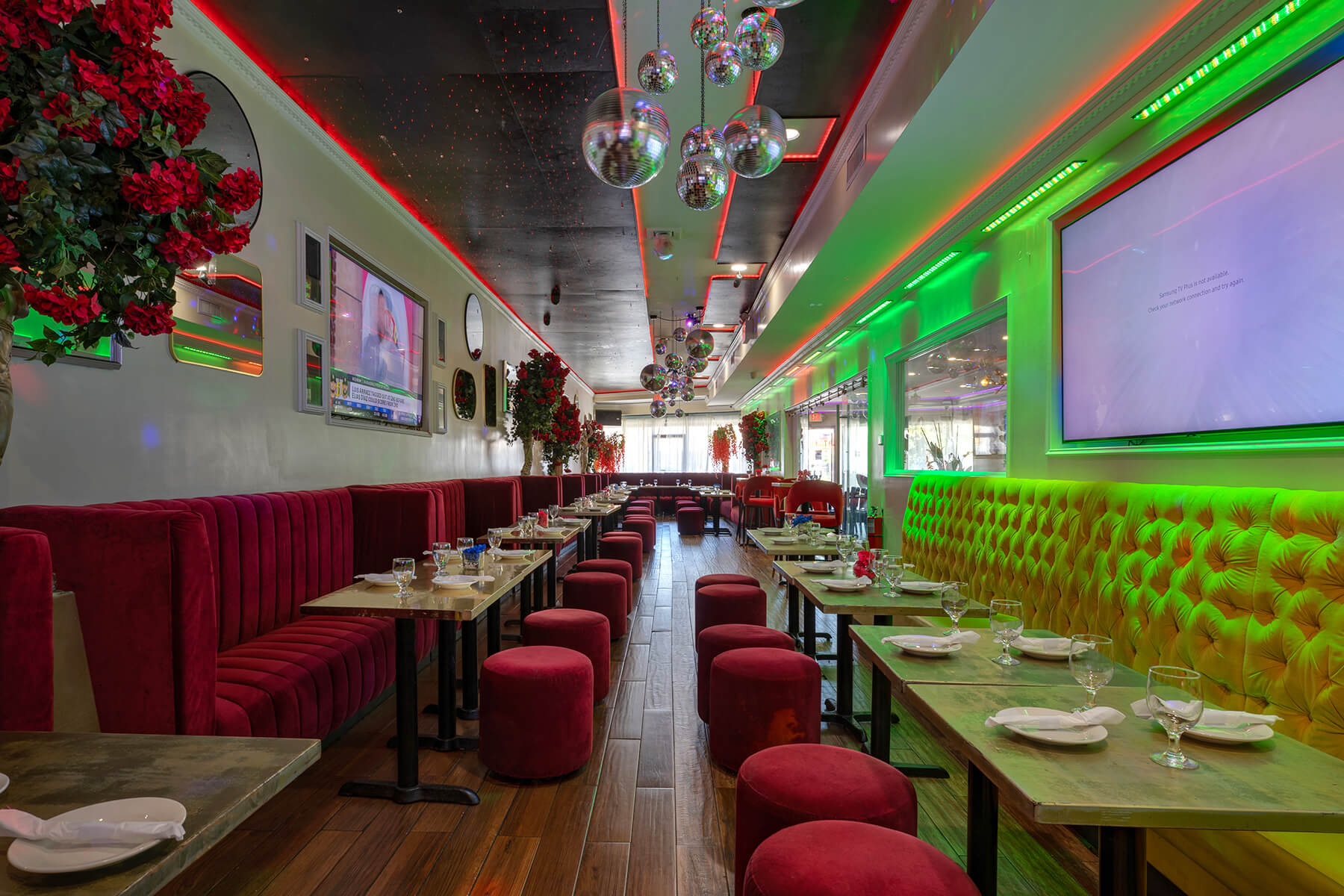 Interior of a modern restaurant with red and yellow tufted seating, small tables set with glasses and plates, disco balls hanging from the ceiling, and green LED lighting on the right wall.