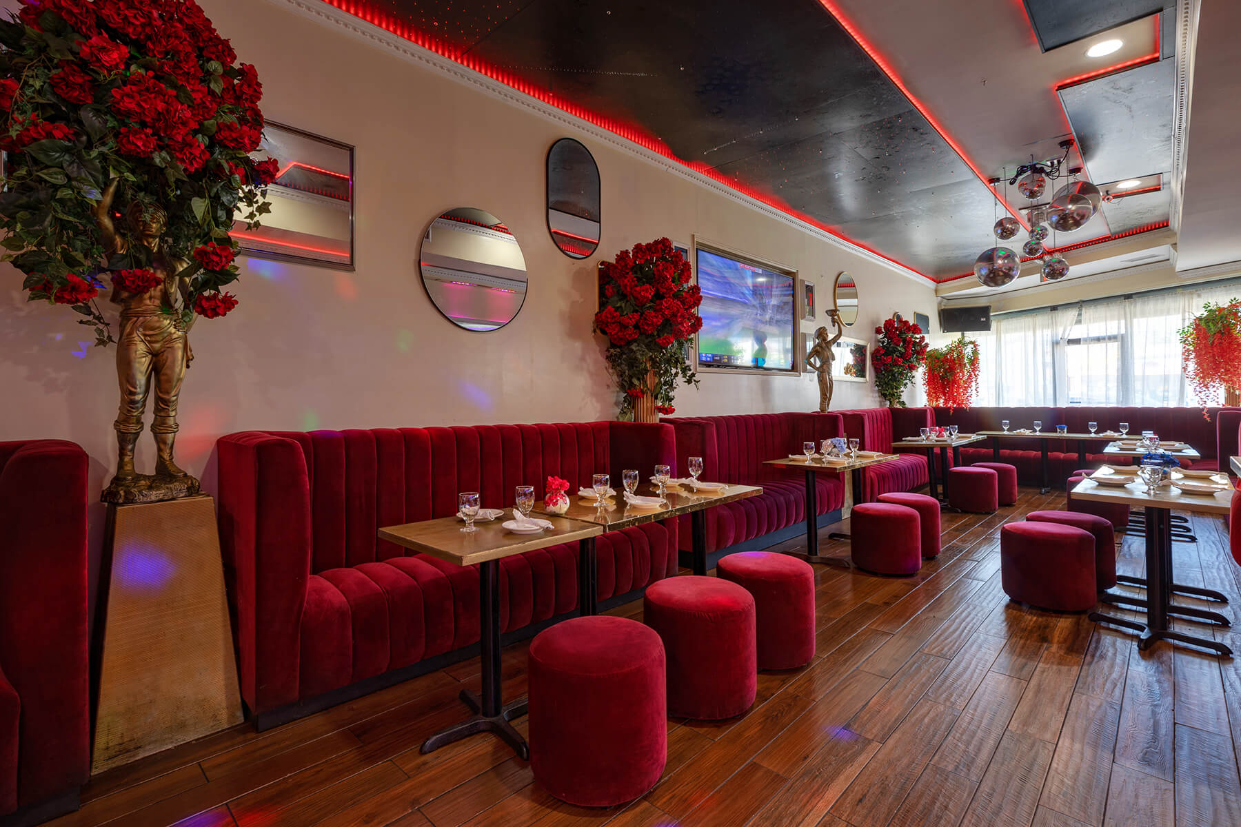 Modern lounge with deep red velvet seating, small tables set with glasses and plates, floral arrangements, statues, and disco balls hanging from the ceiling.