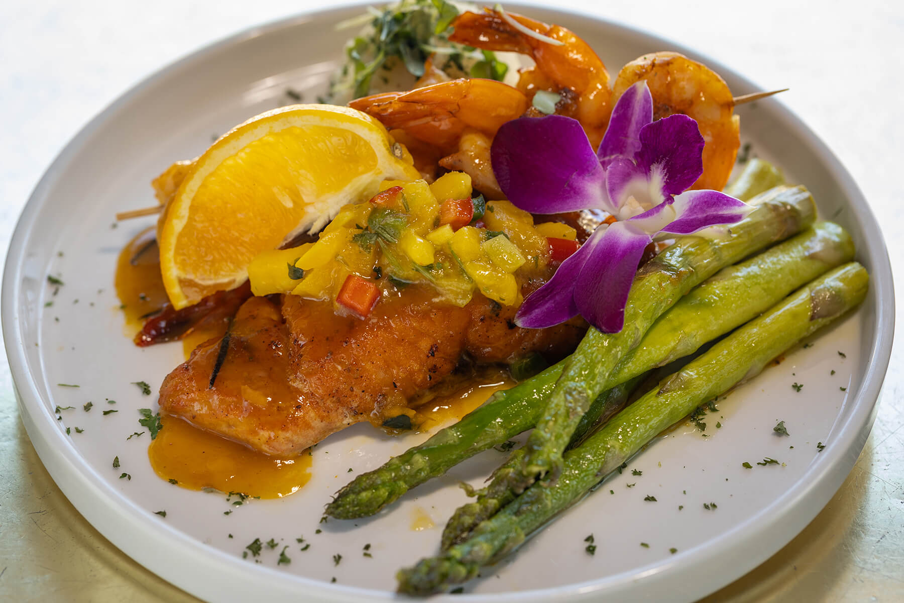 Plate with grilled salmon topped with mango salsa and lemon wedge, shrimp skewer, roasted asparagus, and a purple edible flower garnish.