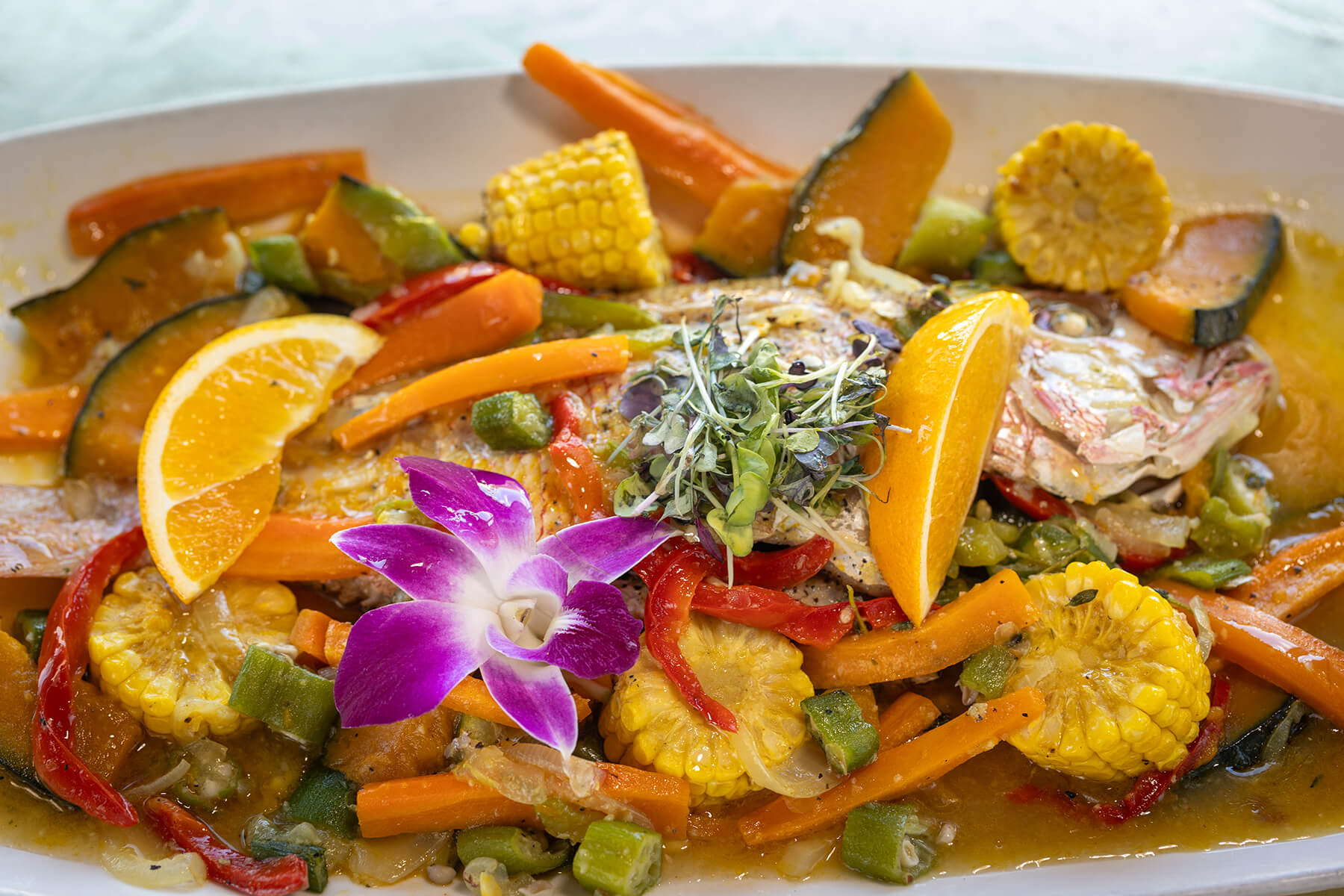 Plated cooked fish garnished with orange slices, microgreens, purple orchid flower, corn, carrots, bell peppers, and squash in sauce.