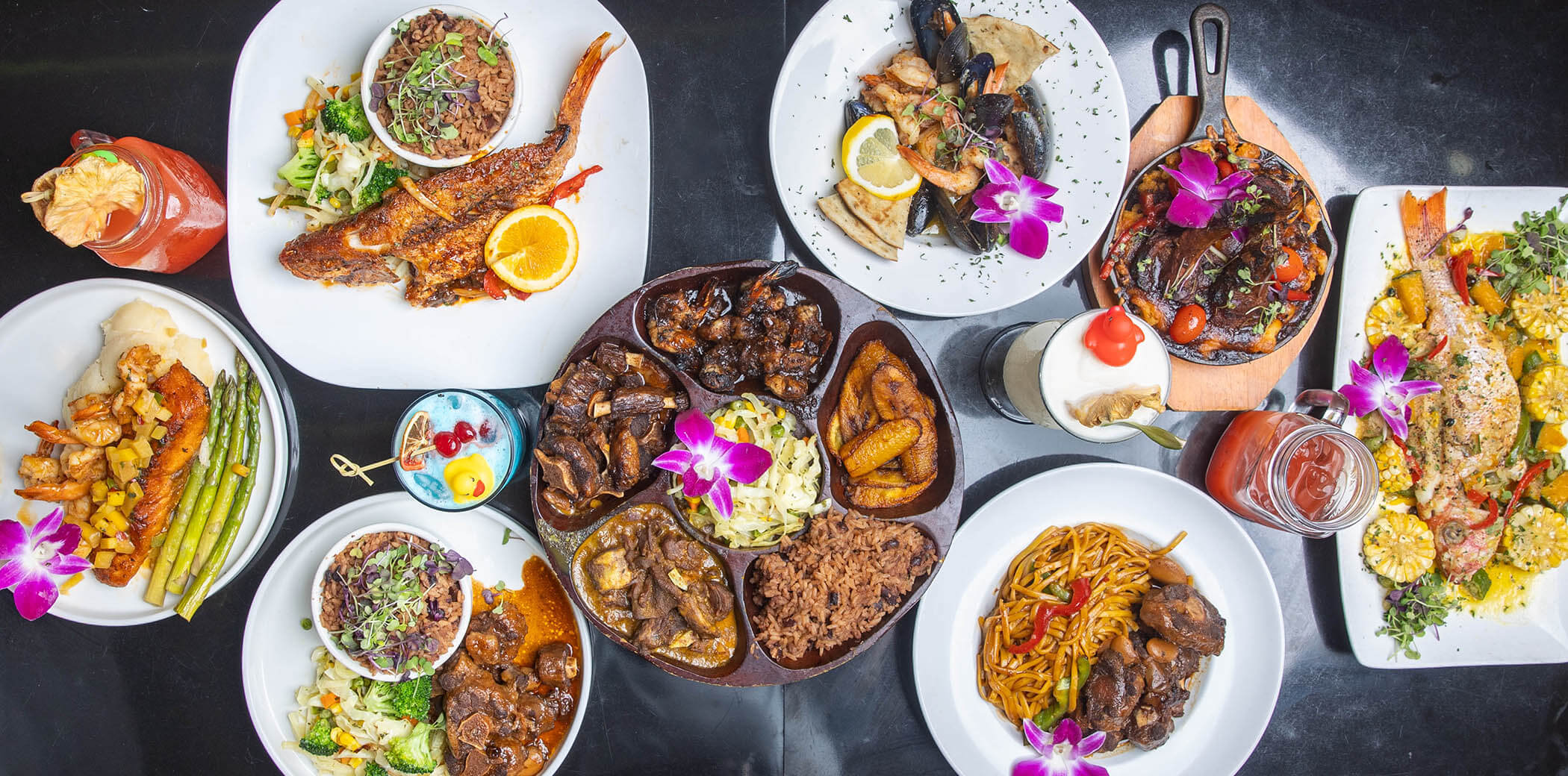 Overhead view of various colorful dishes including grilled fish, shrimp, rice and beans, noodles, plantains, and drinks on a black table, each garnished with purple edible flowers.