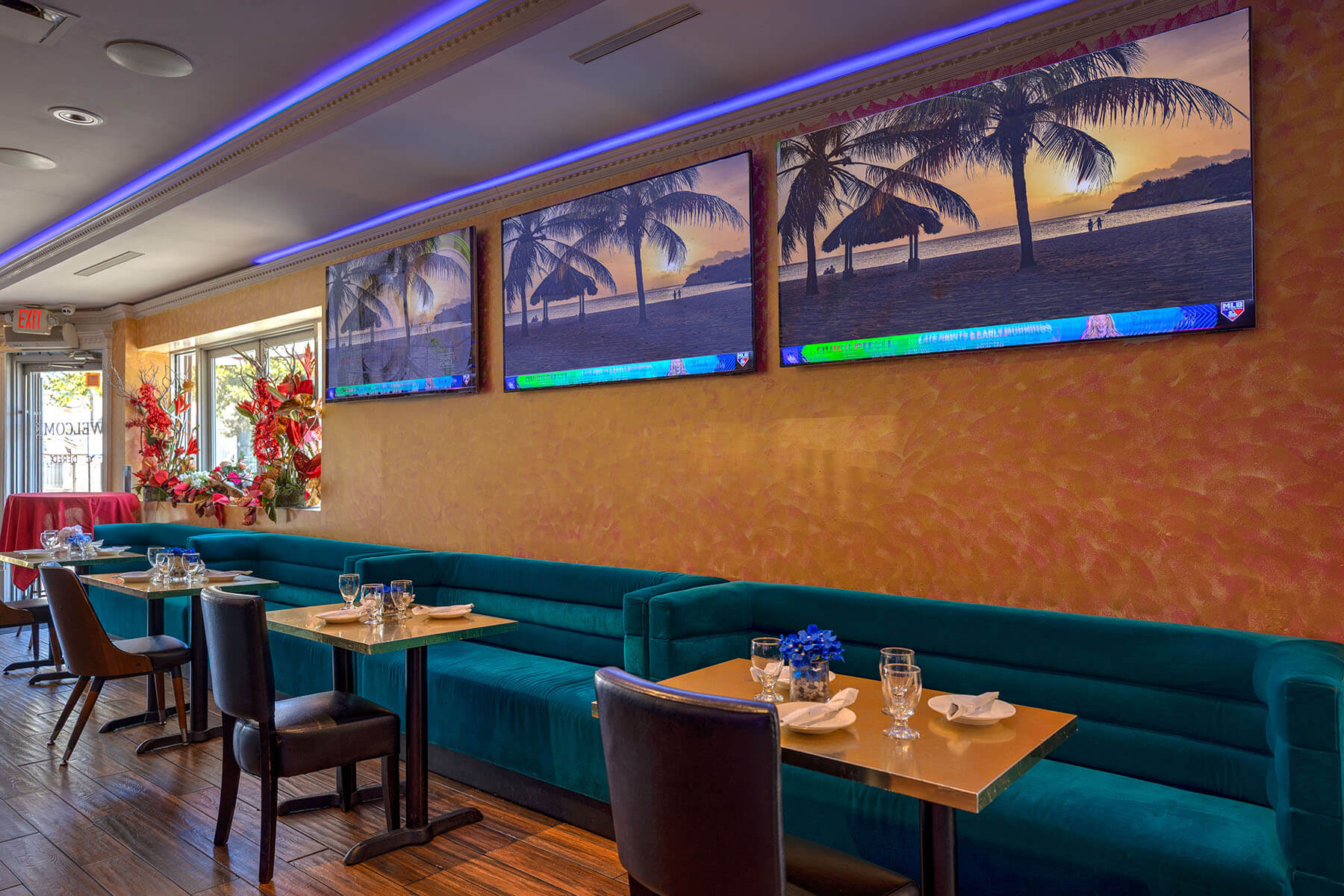 Dining area with teal velvet banquette seating, gold tables set with glasses and napkins, and three large wall TVs showing a tropical beach sunset scene.