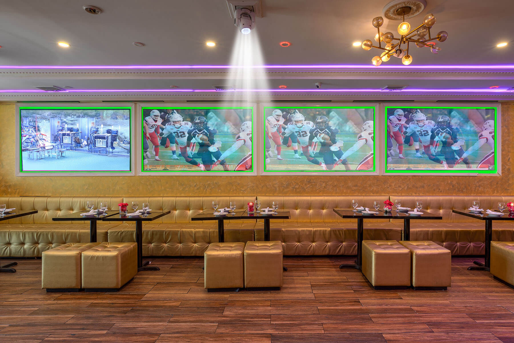 Modern sports bar interior with gold cushioned seating, dark wooden tables set with glassware, and four large TV screens on the wall showing a football game and another image.