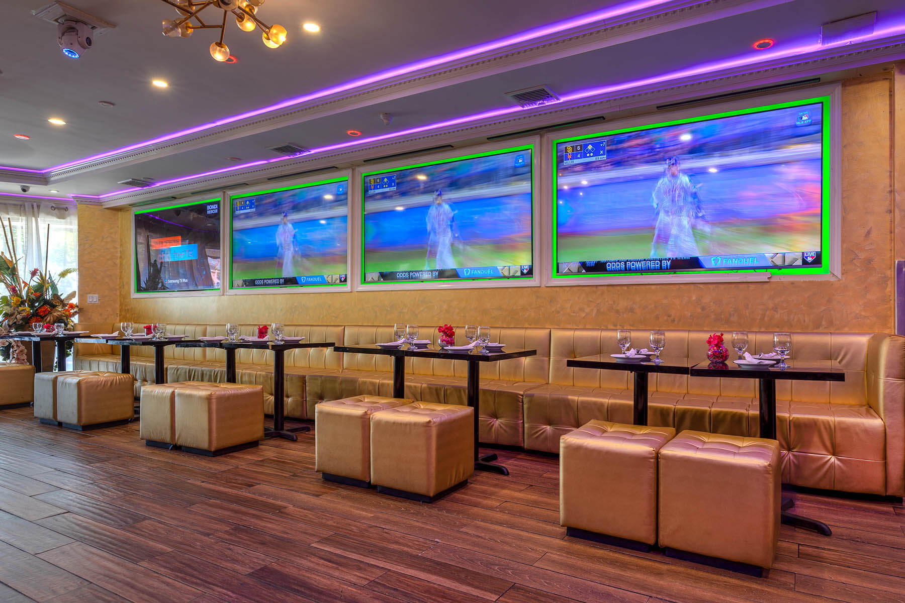 Modern sports bar seating area with tan leather couches and ottomans, black tables set with glasses and napkins, and four large wall-mounted TVs showing a baseball game with purple LED ceiling lighting.