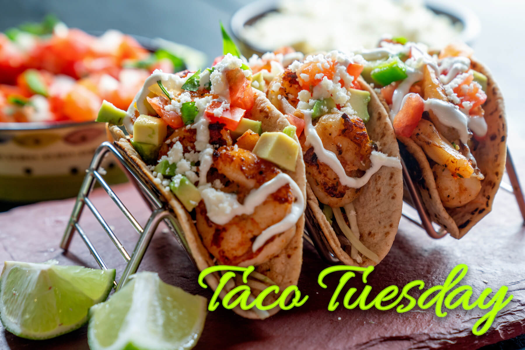 Three shrimp tacos with diced avocado, tomato, cheese, and crema with lime wedges on the side.